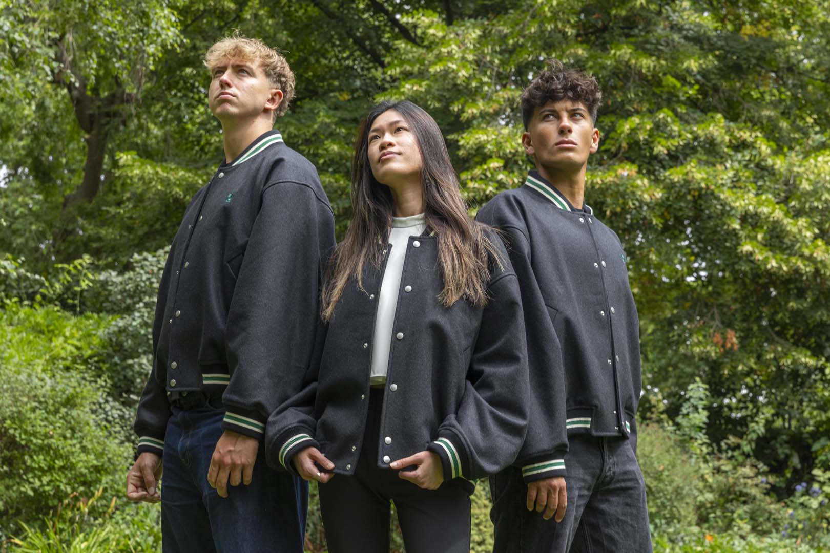 Three people wearing matching dark blue bomber jackets standing outdoors with greenery in the background